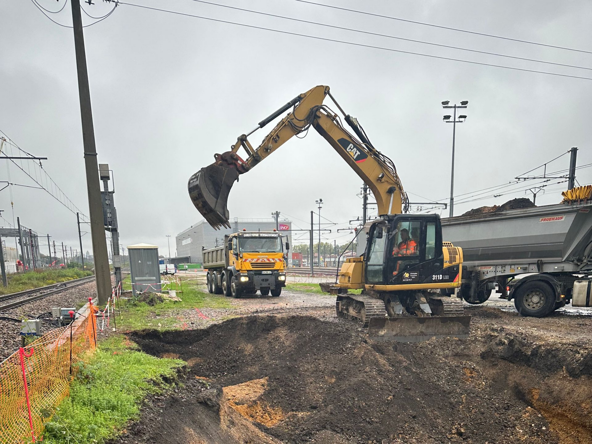 Une excavatrice charge de la terre dans un camion-benne près d'une voie ferrée par temps nuageux.