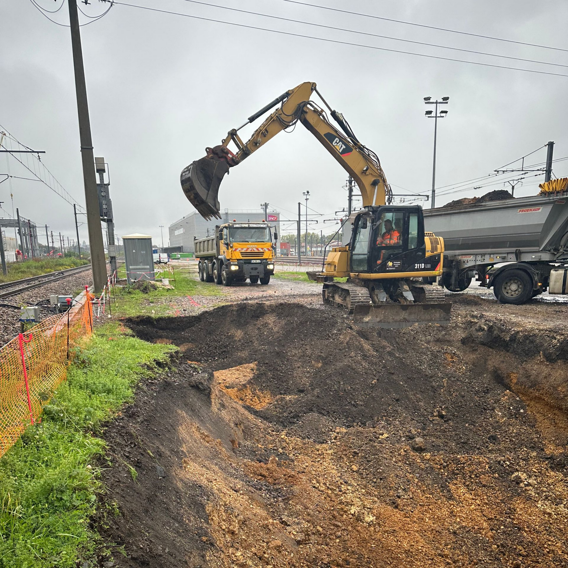 Une excavatrice remplit un camion-benne de terre à côté d'une voie ferrée pendant des travaux de construction, sous un ciel couvert.