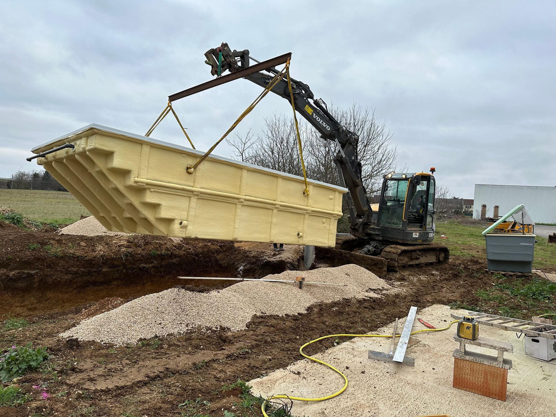 Une pelleteuse soulève une piscine rectangulaire beige et la place dans une zone excavée entourée de gravier et de terre, par temps nuageux.