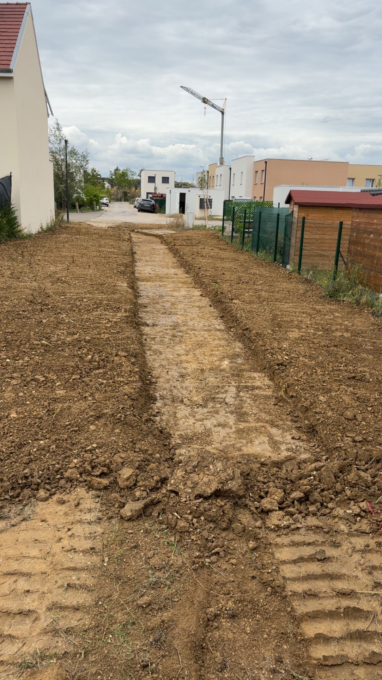 Chemin de terre dans un quartier résidentiel, avec des traces de pneus. Un bâtiment et une grue de chantier sont visibles en arrière-plan.