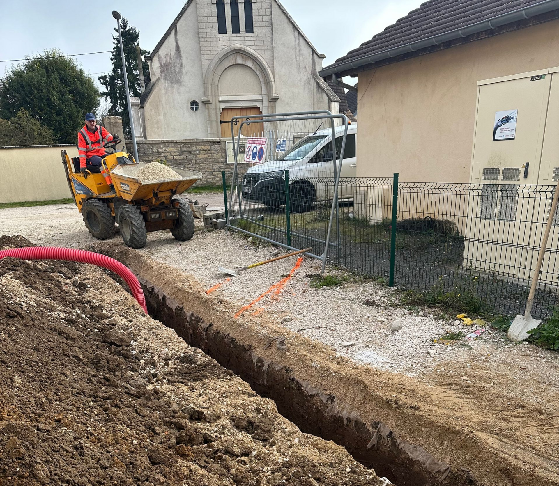 Un homme manœuvre un petit engin de chantier dans une tranchée près d'une église. Un tuyau rouge traverse la tranchée.