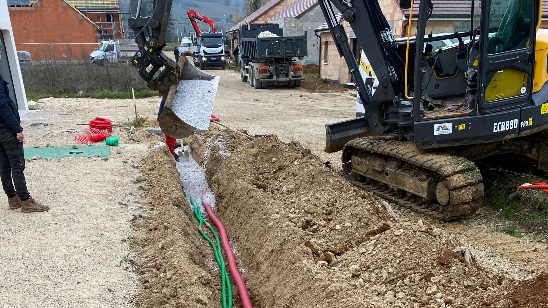 Une pelleteuse enfouit des câbles colorés dans une tranchée sur un chantier. Des camions et une personne se tiennent à proximité.