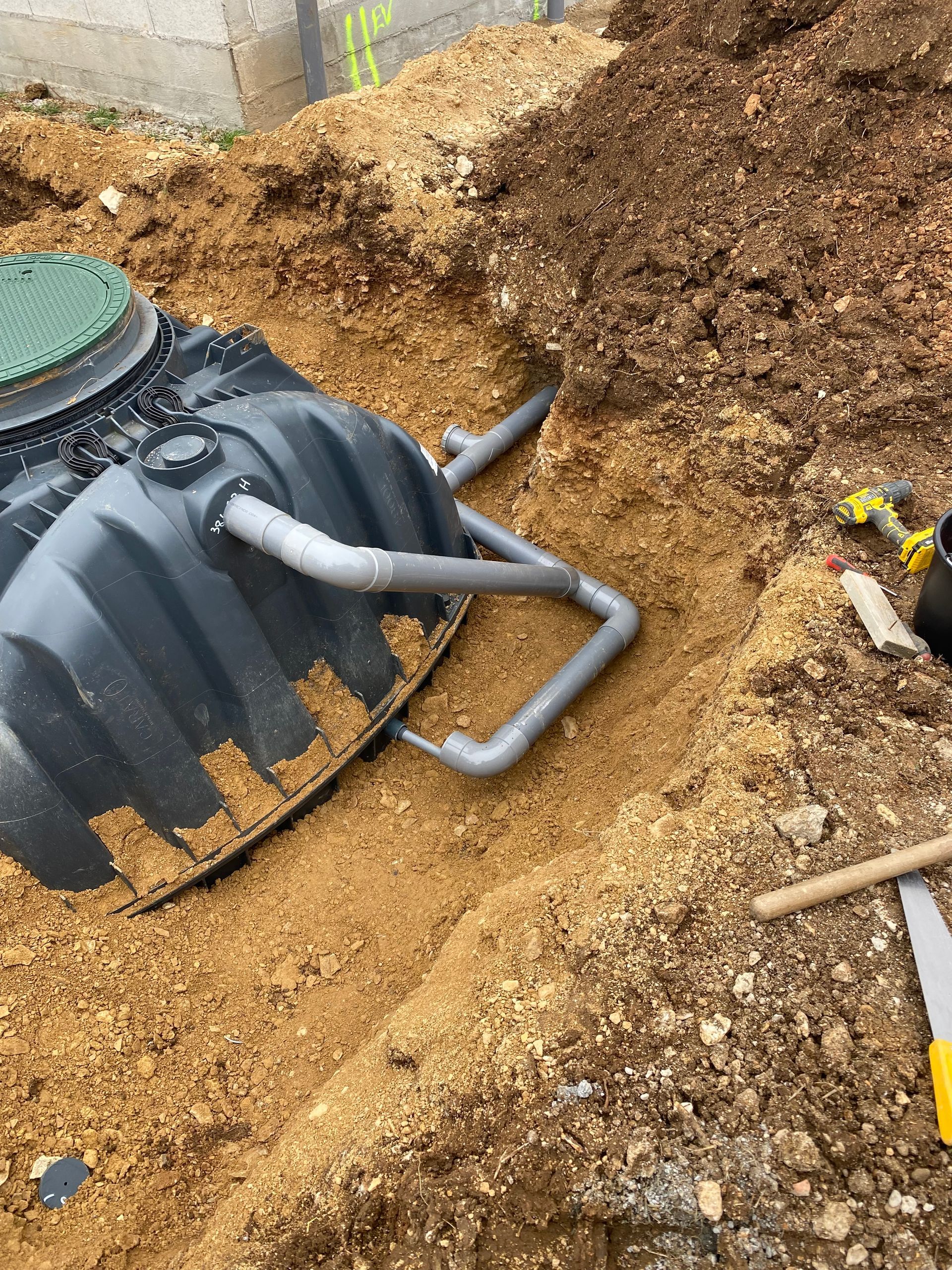 Fosse septique noire avec tuyaux raccordés, dans une tranchée creusée, entourée de terre et d'outils.