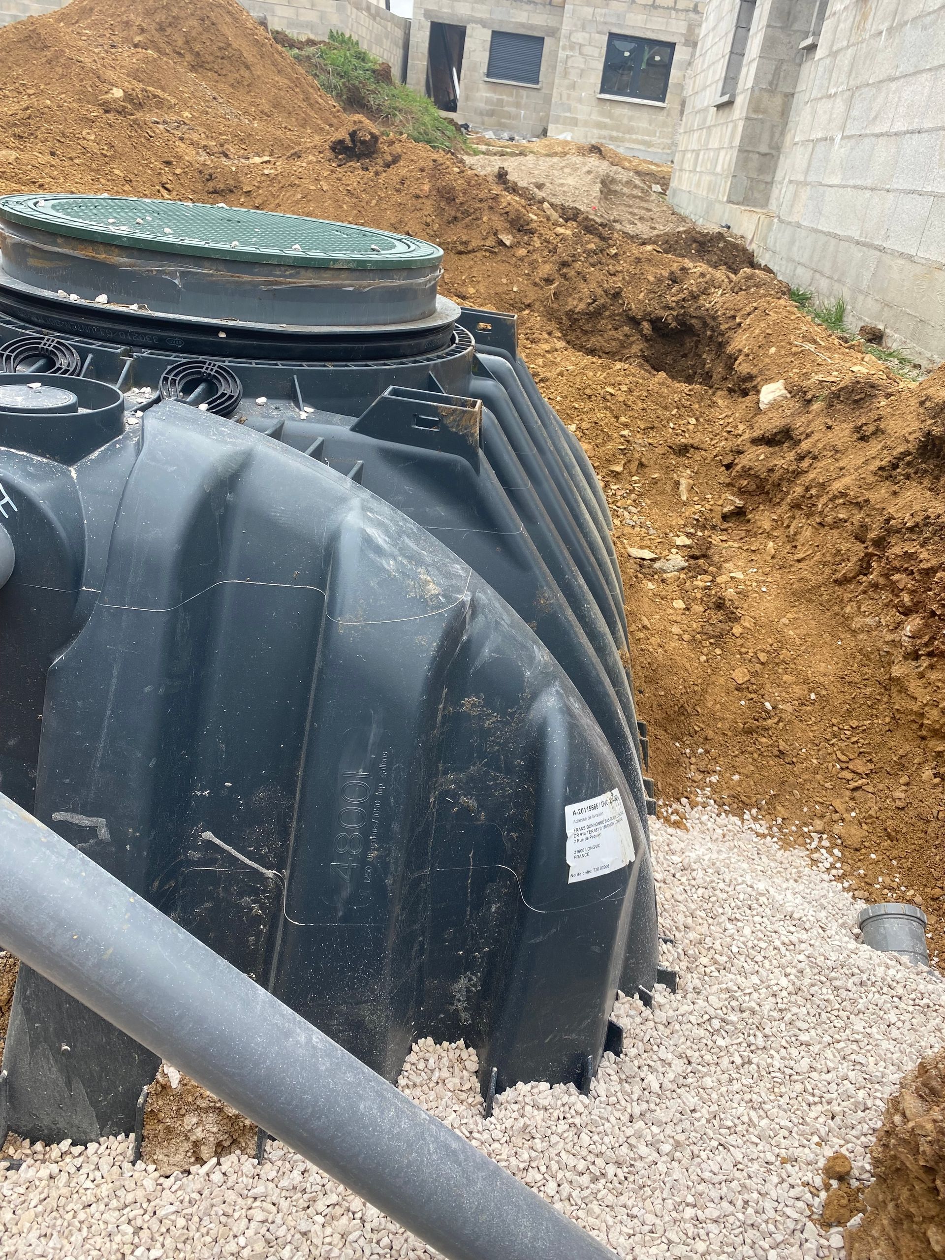 Fosse septique noire dans un trou partiellement creusé, entourée de gravier et de terre, près d'un bâtiment.