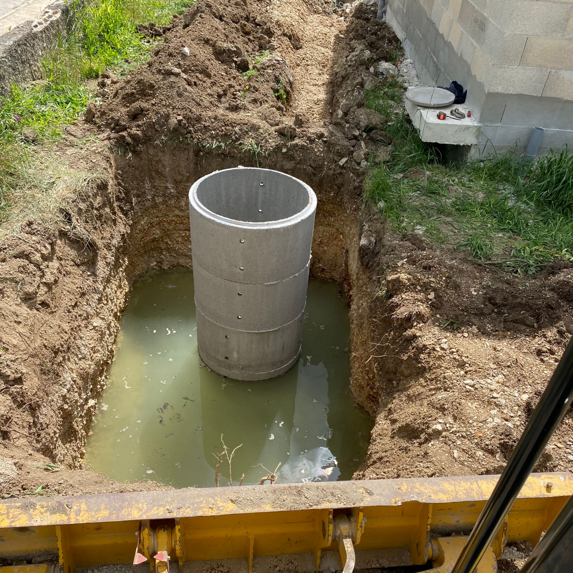 Cylindre de béton dans une excavation remplie d'eau. Des parois de terre l'entourent, à proximité d'un bâtiment.
