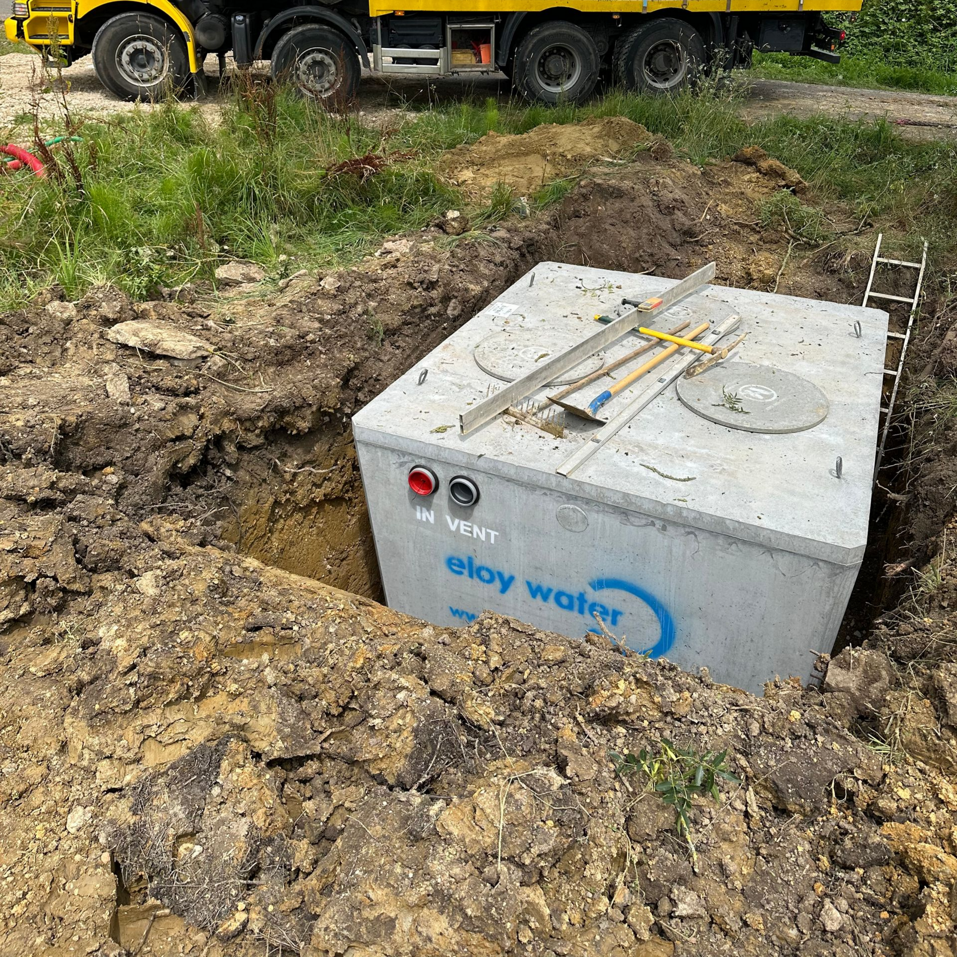 Réservoir en béton portant l'inscription « eau eloy » dans une tranchée de chantier, avec un camion en arrière-plan.