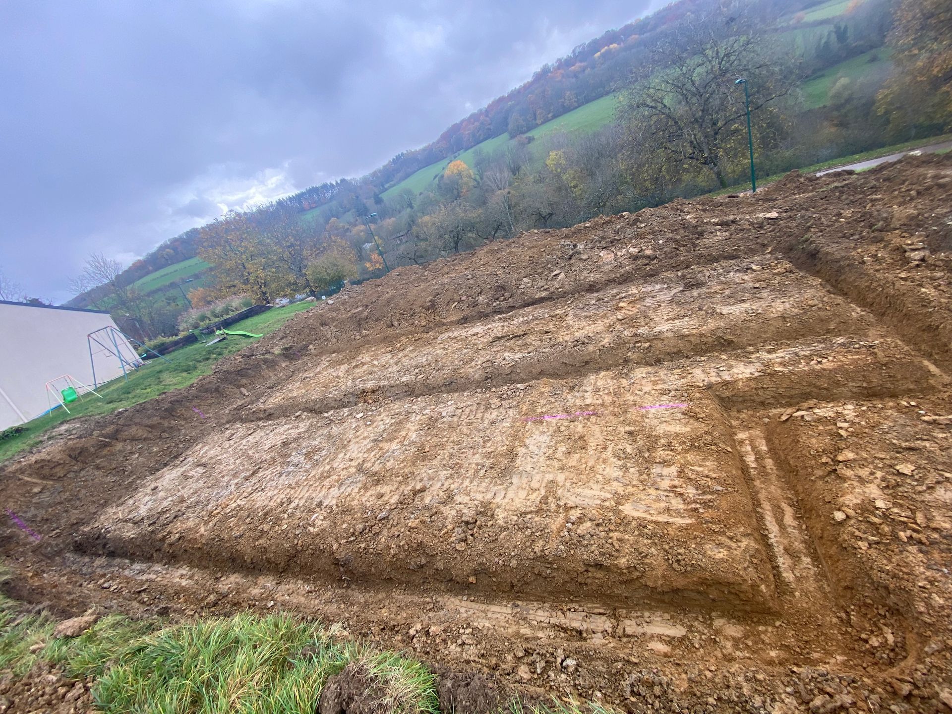 Fondation rectangulaire creusée à flanc de colline, aux tons terreux, avec une zone herbeuse et de la végétation en arrière-plan.