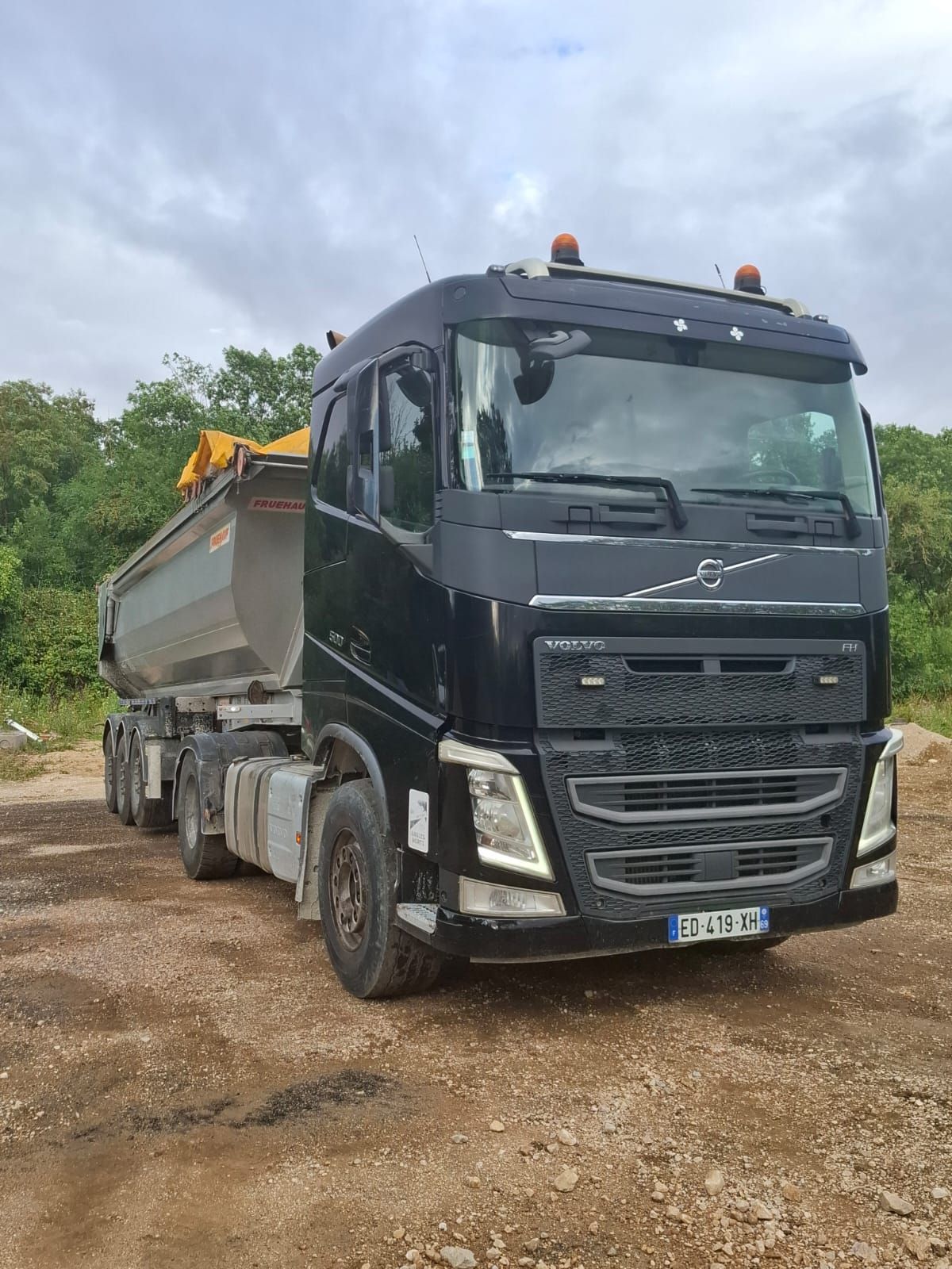 Un semi-remorque Volvo noir avec une benne basculante argentée est stationné sur du gravier, sous un ciel nuageux.