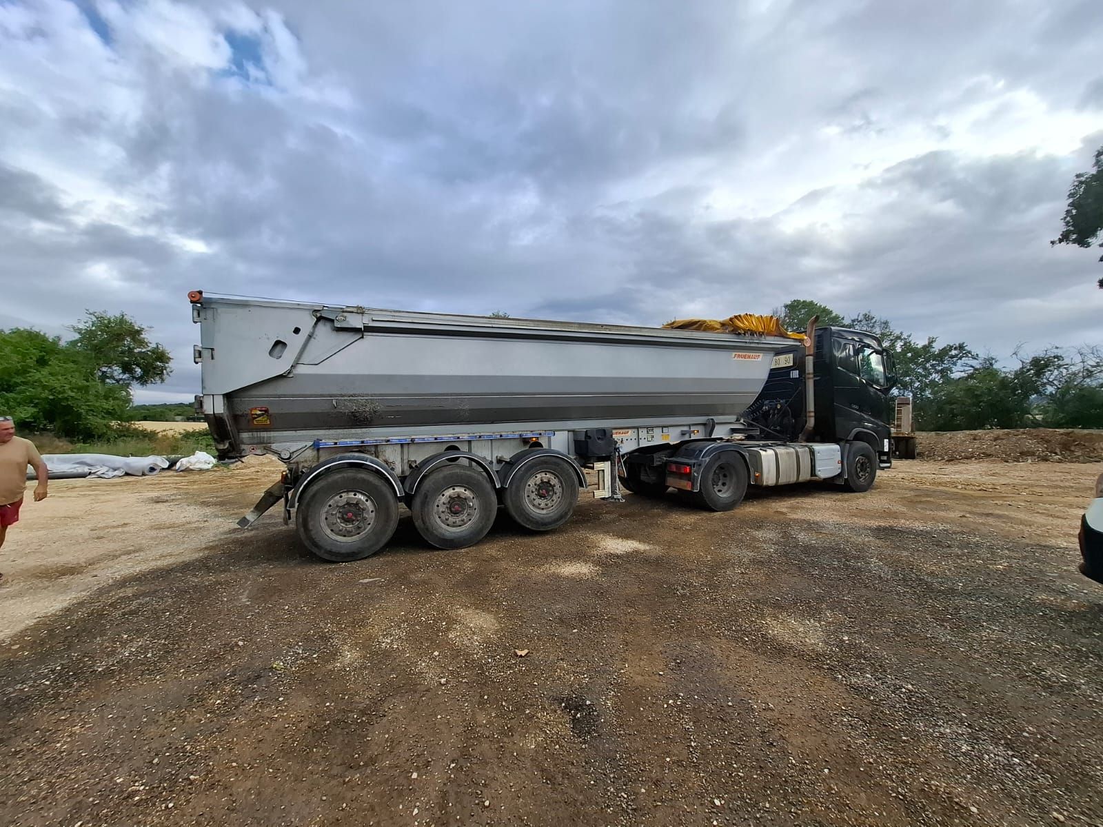 Un gros camion-benne gris sur du gravier, prêt à décharger. Ciel nuageux en arrière-plan, une personne à gauche.