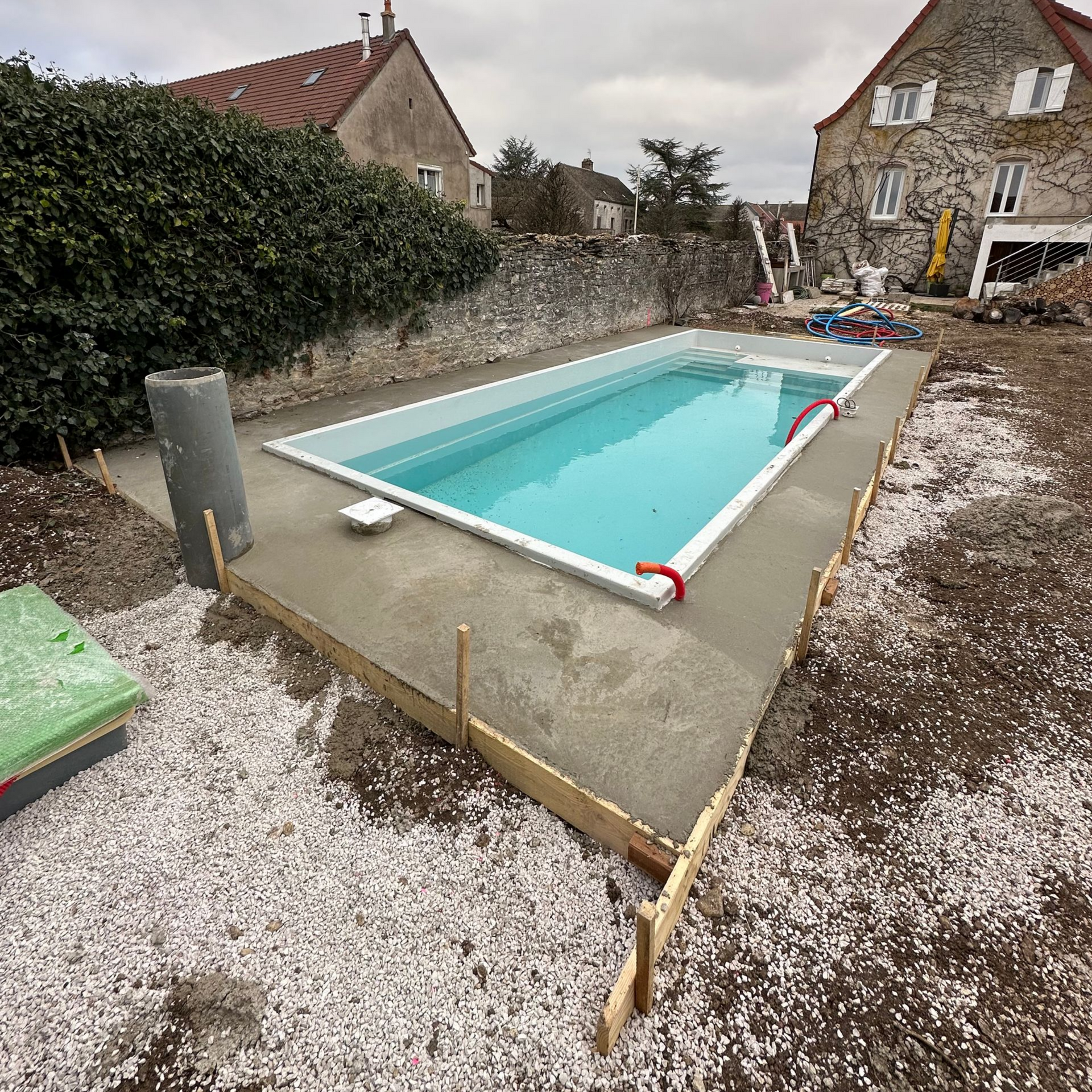 Piscine rectangulaire en construction, bordure en béton, sol en gravier, maisons en pierre de deux étages en arrière-plan.