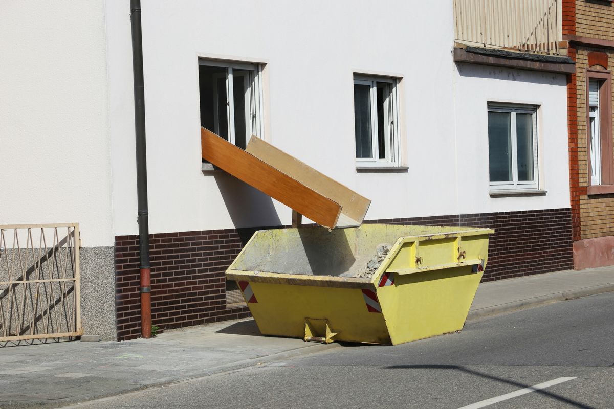 Vor einem weißen Gebäude steht ein gelber Müllcontainer.