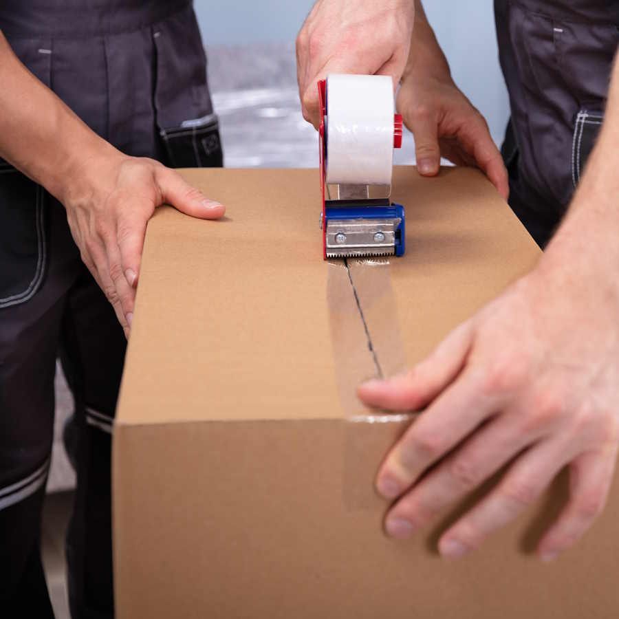 Ein Umzugshelfer klebt mit Paketband einen Umzugskarton zu. Er und ein weiterer Umzugshelfer halten dabei den Umzugskarton fest. Wir sind Ihre zuverlässige Umzugsfirma aus Zürich.