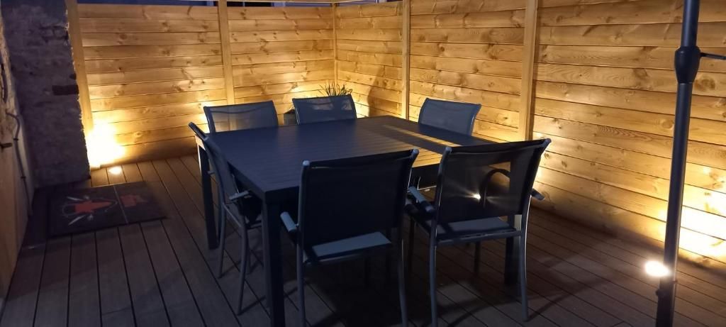 Espace repas extérieur avec table et chaises sur une terrasse en bois, éclairé par des spots contre une clôture en bois.