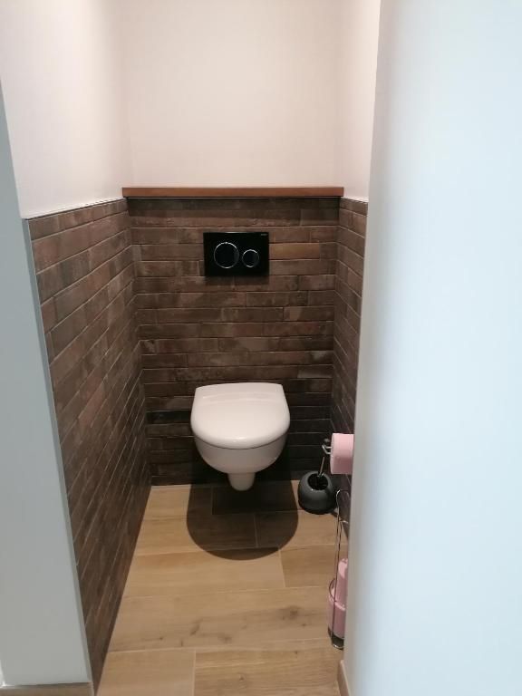 Petite salle de bains avec carrelage imitation brique marron sur les murs, parquet, toilettes blanches et plaque de chasse d'eau.