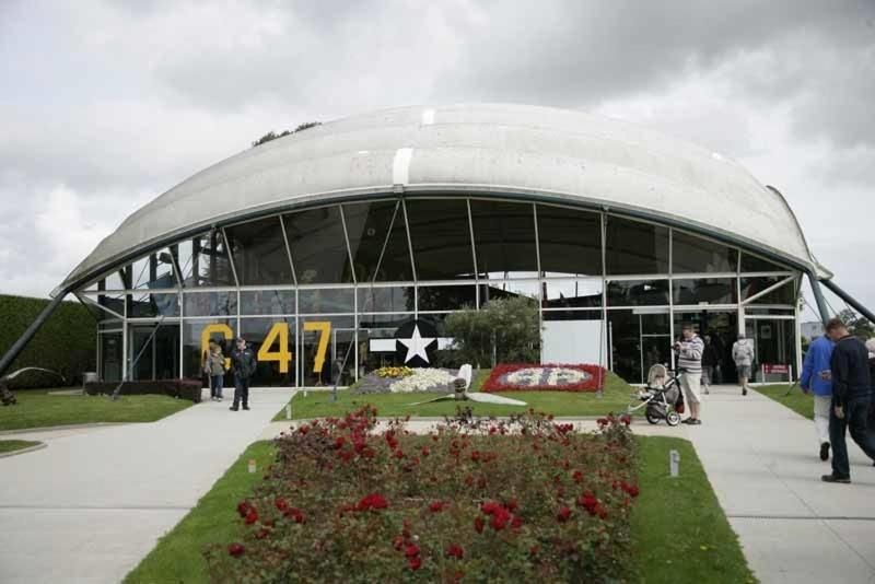 Grand bâtiment blanc au toit voûté avec un panneau « C47 » et des gens à l'extérieur, un jardin devant.