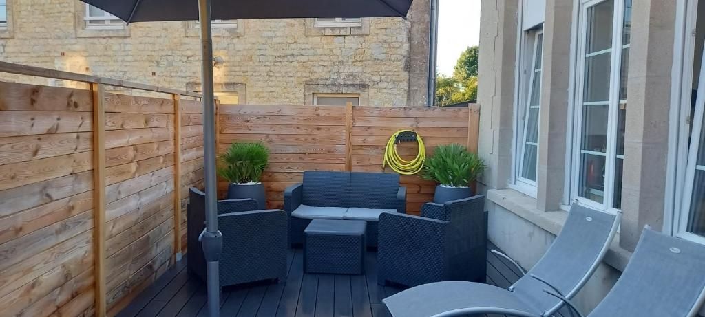 Terrasse extérieure avec sièges, parasol, clôture en bois et bâtiment en arrière-plan.