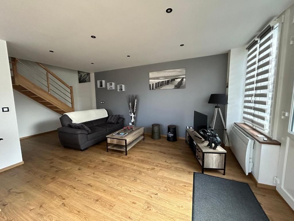 Salon avec canapé gris, parquet et escalier. Mur gris orné d'œuvres d'art et d'une télévision.