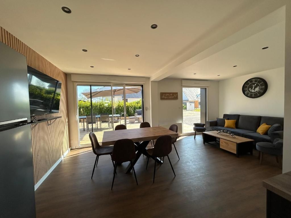 Salon moderne avec coin repas et accès extérieur. Table en bois foncé, canapé gris, murs clairs.