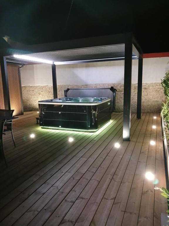 Bain à remous sur une terrasse en bois sous une pergola noire, éclairé par des spots au sol, la nuit.
