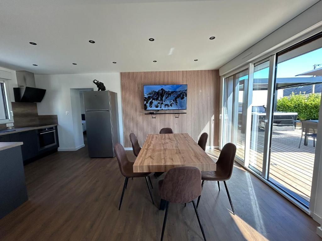 Salle à manger moderne avec table et chaises, télévision, cuisine et portes coulissantes donnant sur une terrasse.