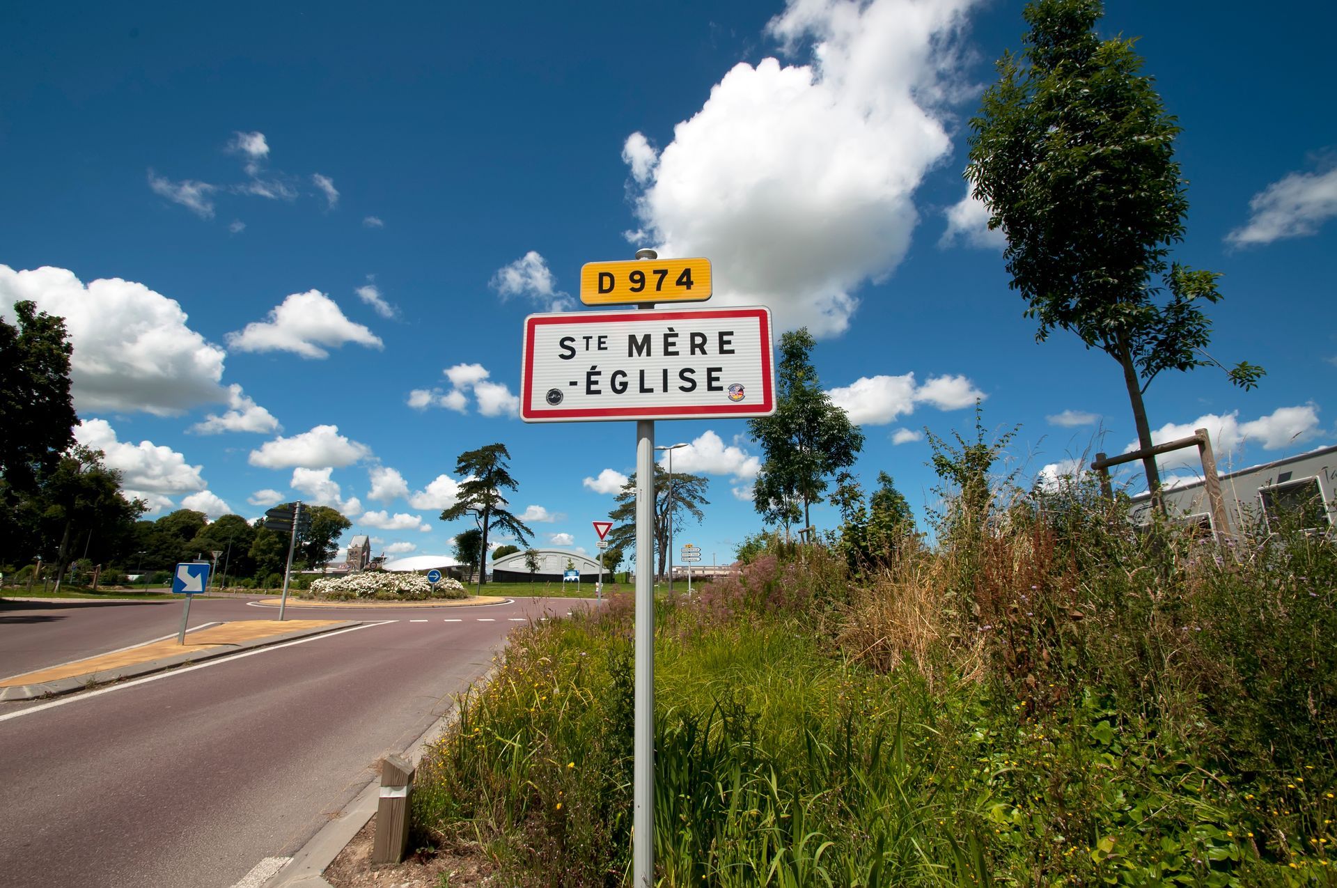 Panneau de Sainte-Mère-Église, France, sous un ciel bleu avec des nuages.