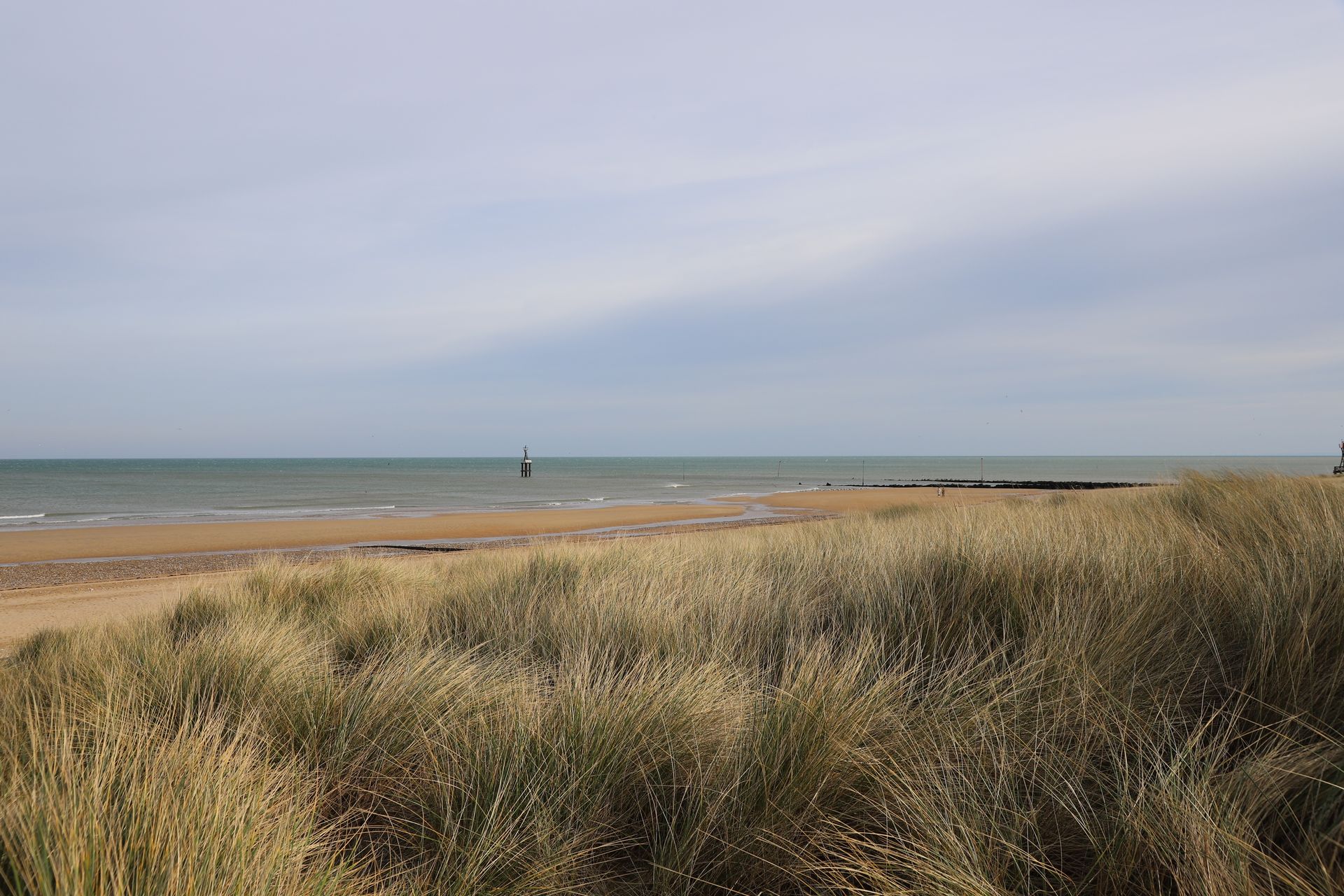 Dunes herbeuses sur une plage, menant à la mer sous un ciel nuageux.