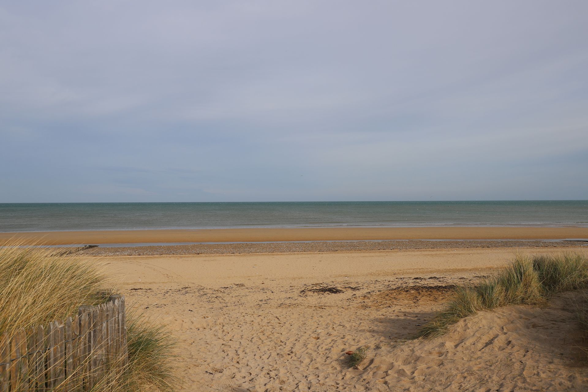 Scène de plage : sable, mer et ciel nuageux. L'herbe des dunes et la clôture en bois encadrent le premier plan sablonneux.