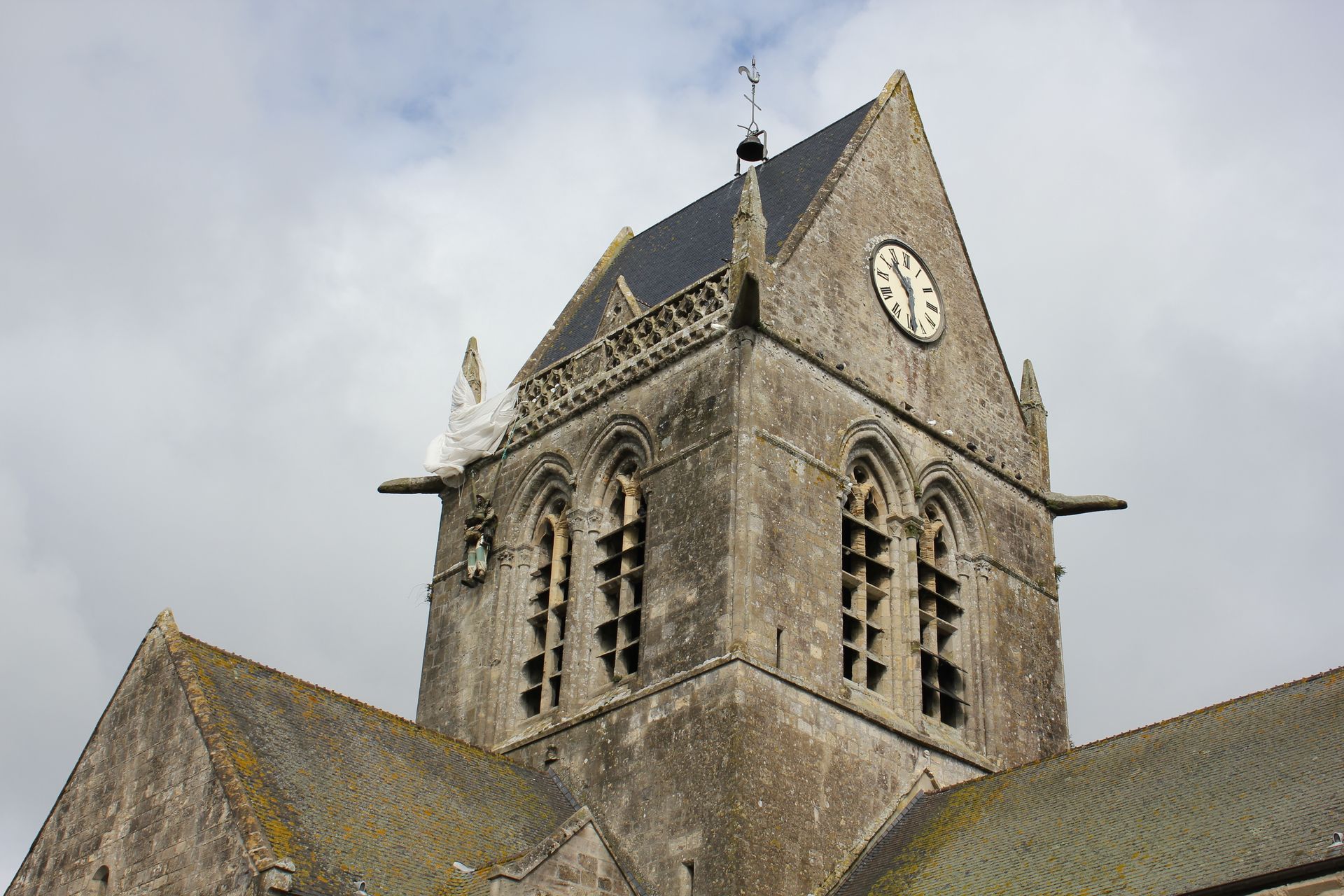 Clocher d'église avec horloge, façade en pierre et toit sombre sur fond de ciel nuageux.