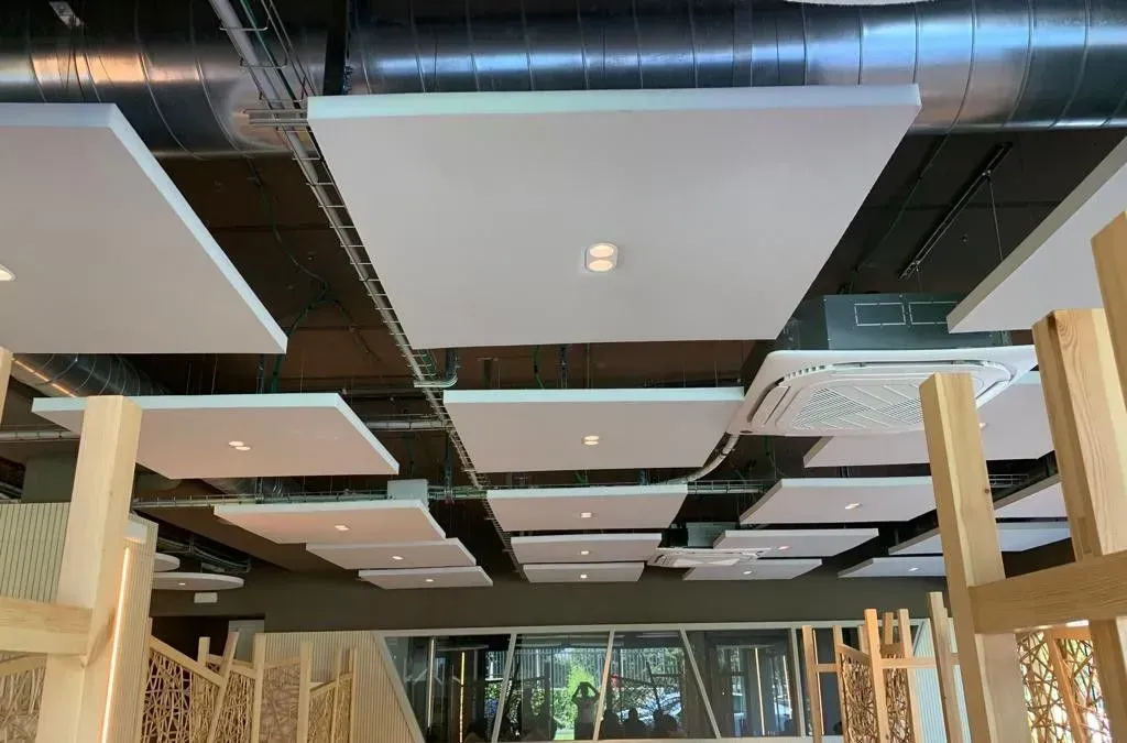 Paneles acústicos blancos suspendidos de un techo oscuro en un espacio comercial, cerca de vigas de madera.