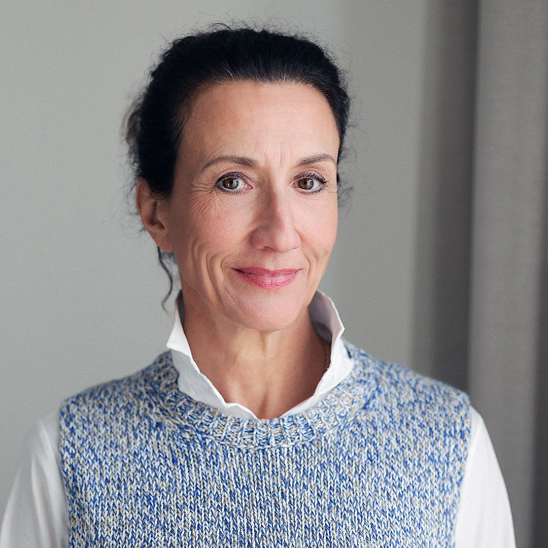 Eine Frau mit dunklen Haaren, die eine blaue Strickweste und ein weißes Hemd trägt, lächelt.