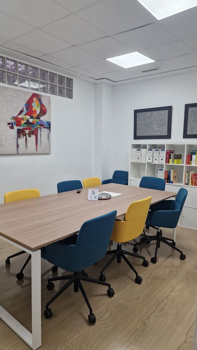 Pequeña sala de reuniones con una mesa de madera, sillas azules y amarillas, cuadros en las paredes y un tablón de anuncios.