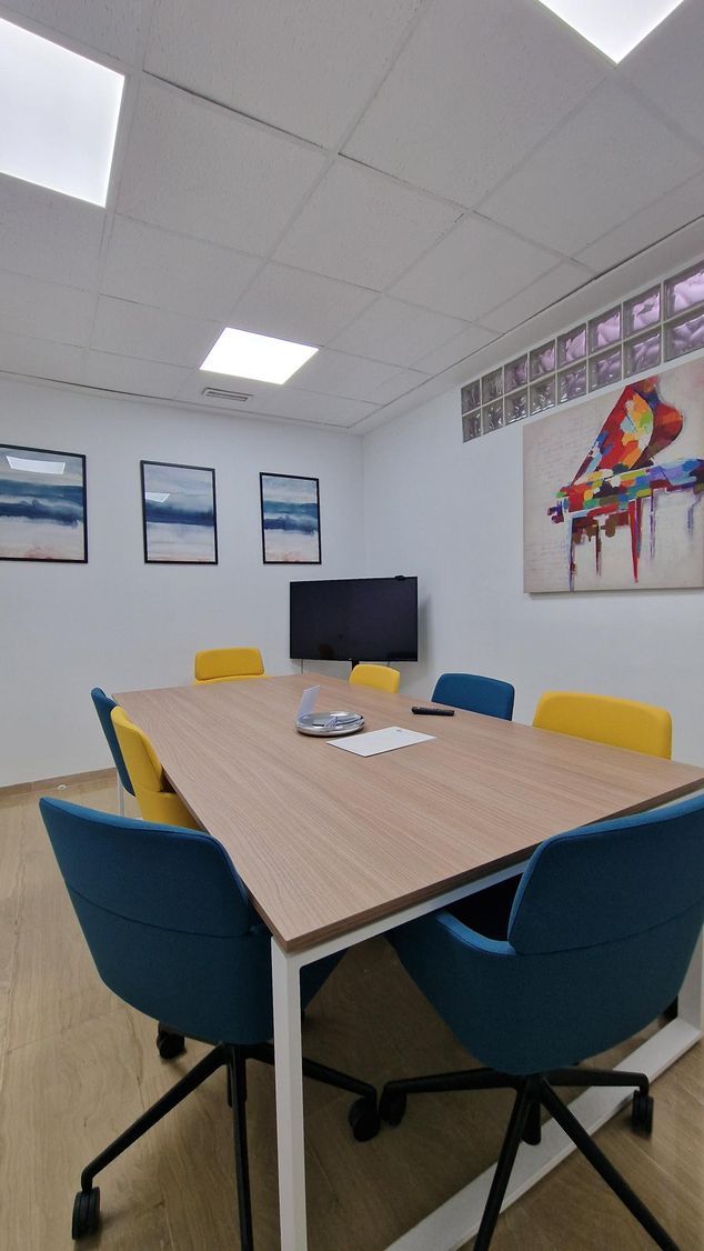 Sala de conferencias vacía con una mesa de madera, sillas azules y amarillas, cuadros en la pared 