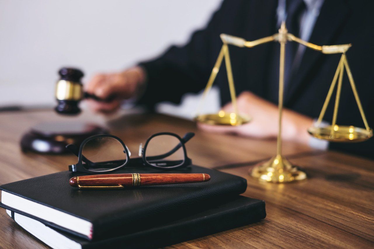 Libro de leyes, gafas y mazo sobre un escritorio con balanzas de la justicia al fondo borroso.