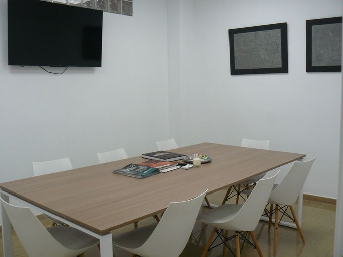 Sala de conferencias con mesa de madera, sillas blancas, televisor montado en la pared y cuadros enmarcados.