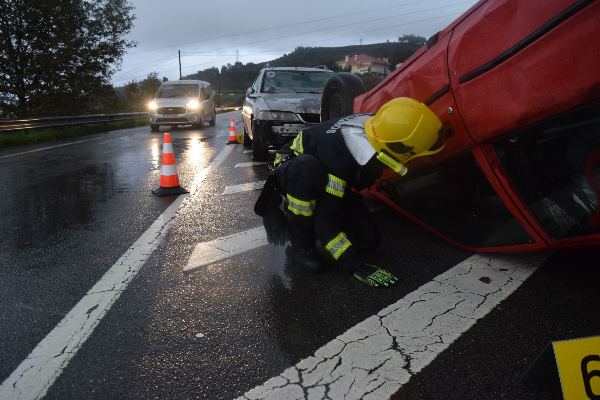 Un bombero se arrodilla junto a un vehículo rojo volcado en una carretera mojada al anochecer