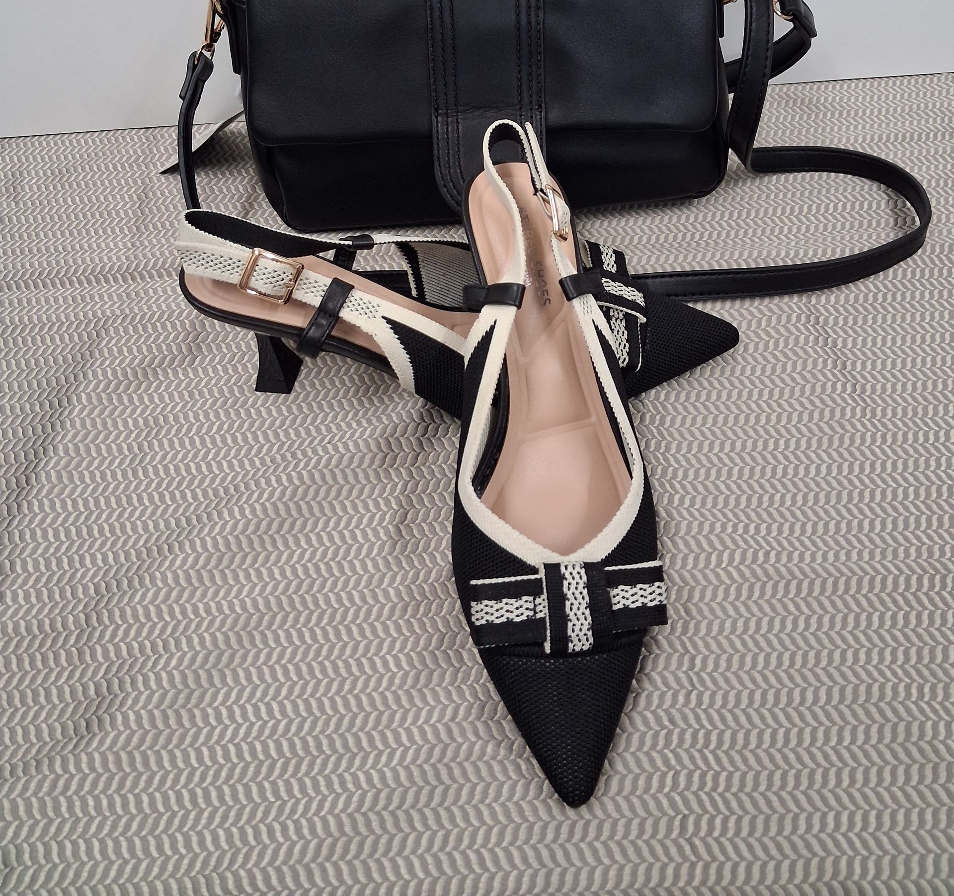 Zapatos planos negros de punta con tira trasera y ribete blanco junto a un bolso negro sobre una superficie texturizada gris.