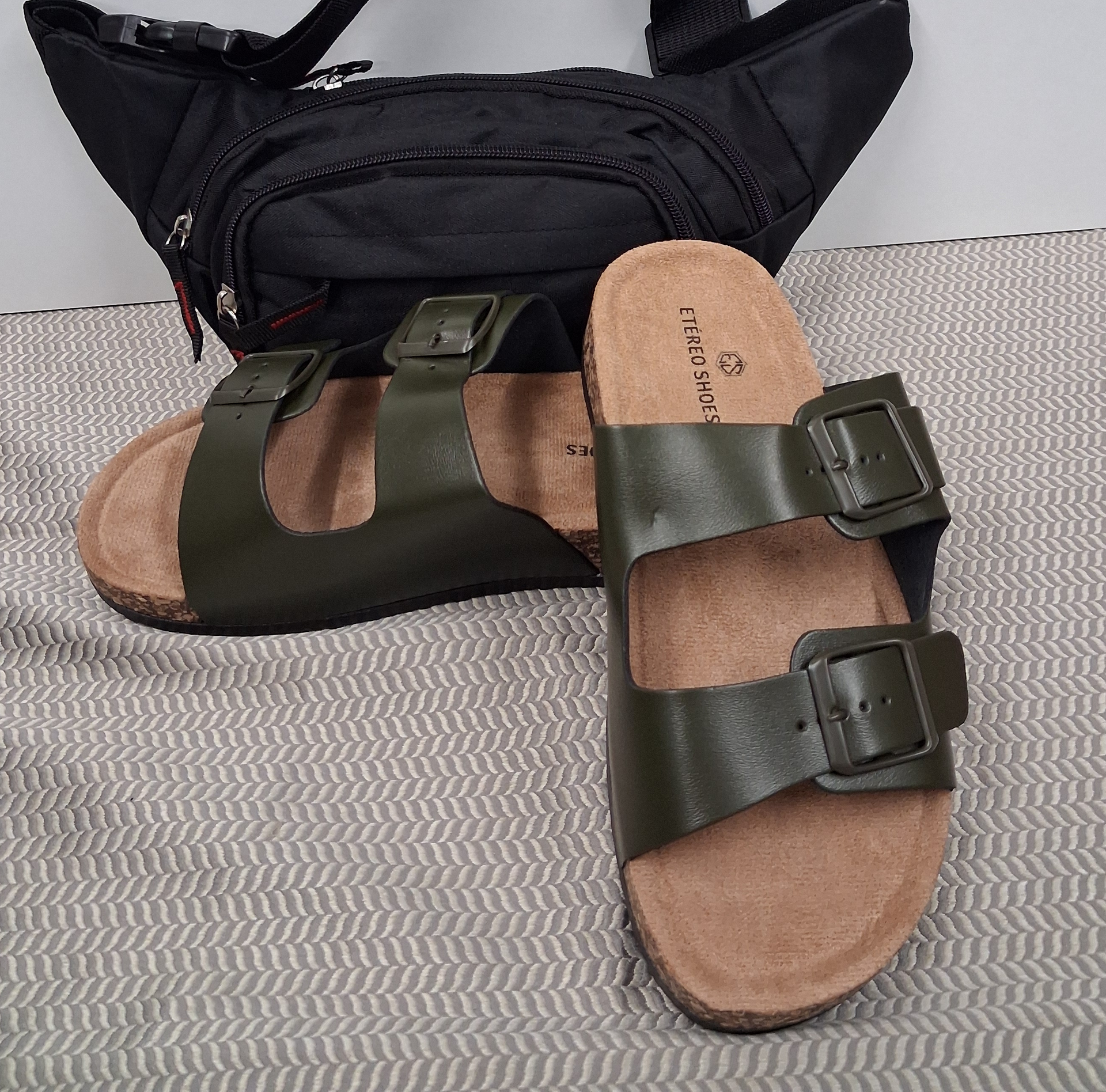 Riñonera negra detrás de sandalias de doble hebilla color oliva y tostado sobre una superficie texturizada gris.