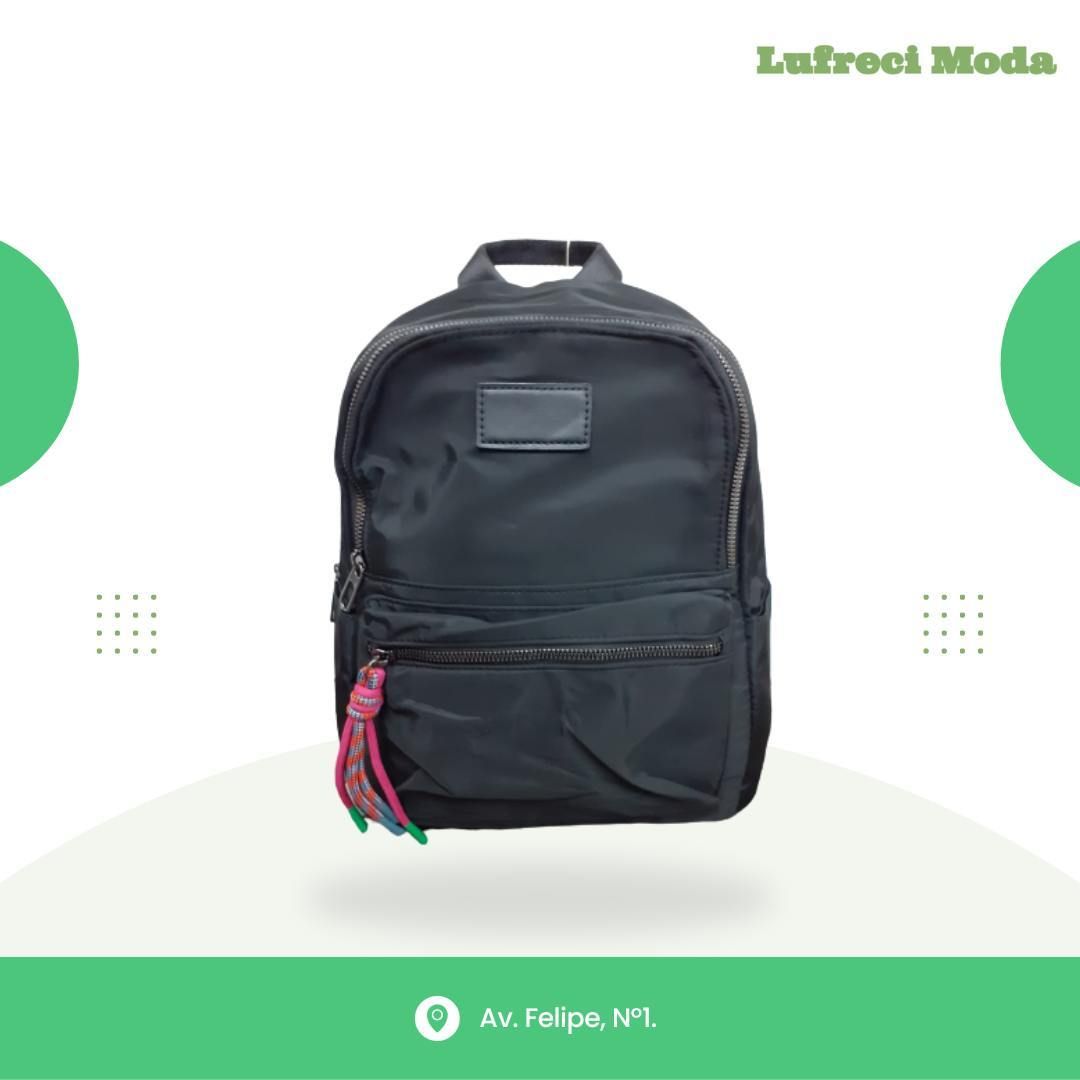 Mochila negra con borla multicolor en la cremallera, centrada sobre un fondo claro con acentos decorativos verdes.