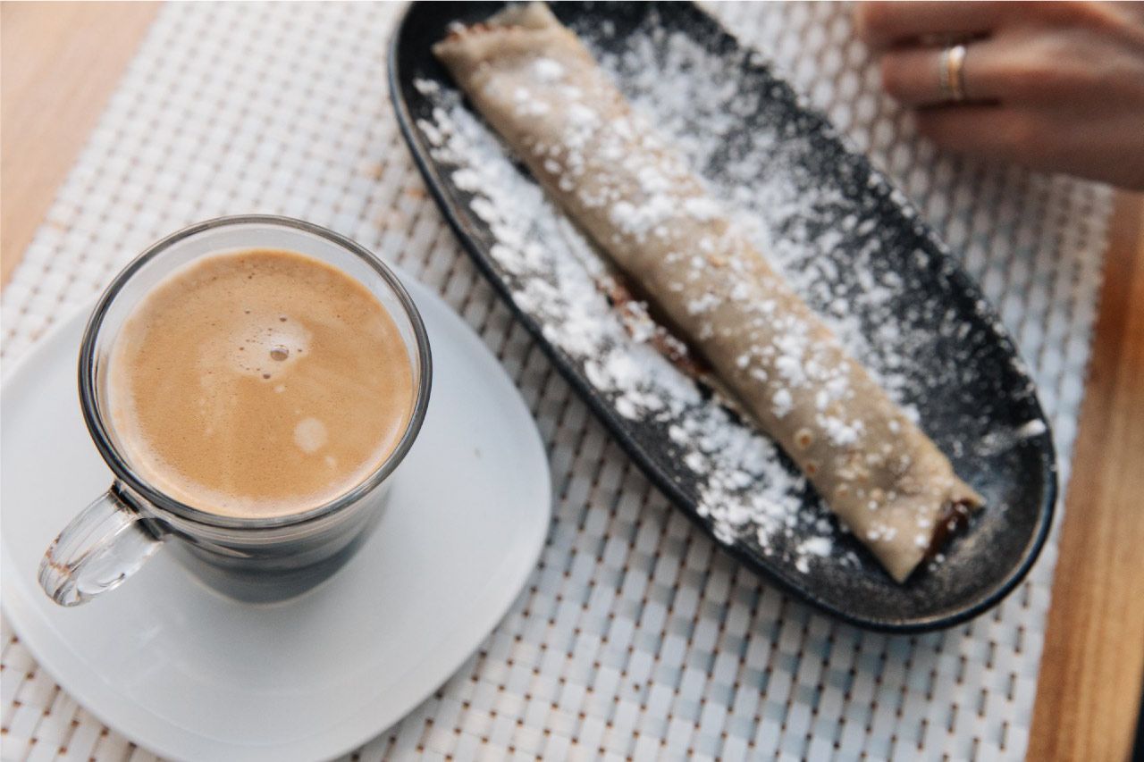 Café en taza de vidrio junto a un pastel enrollado con azúcar glas sobre un plato negro.