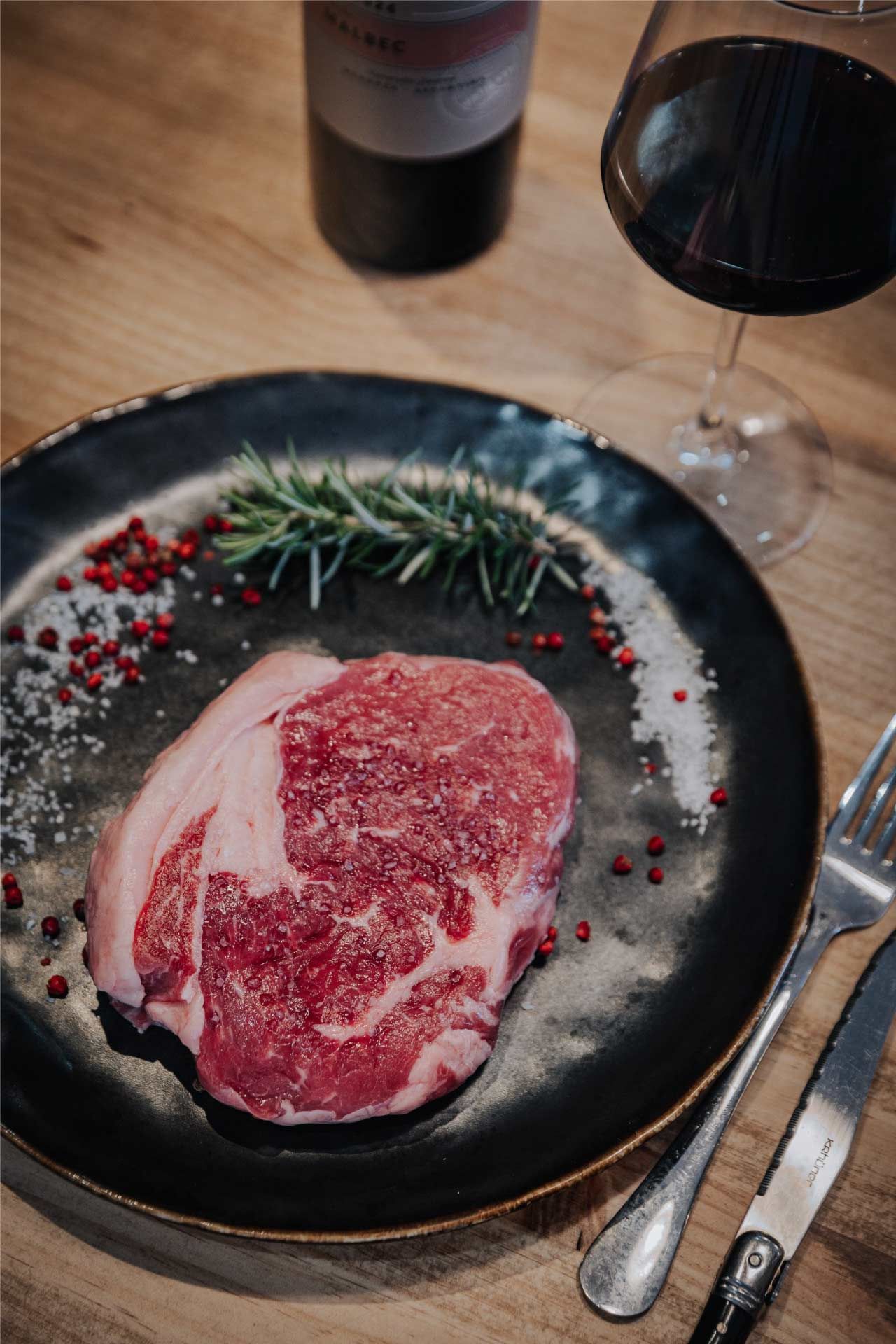 Filete crudo en un plato con condimentos, una copa de vino tinto y una botella de vino sobre una mesa de madera.