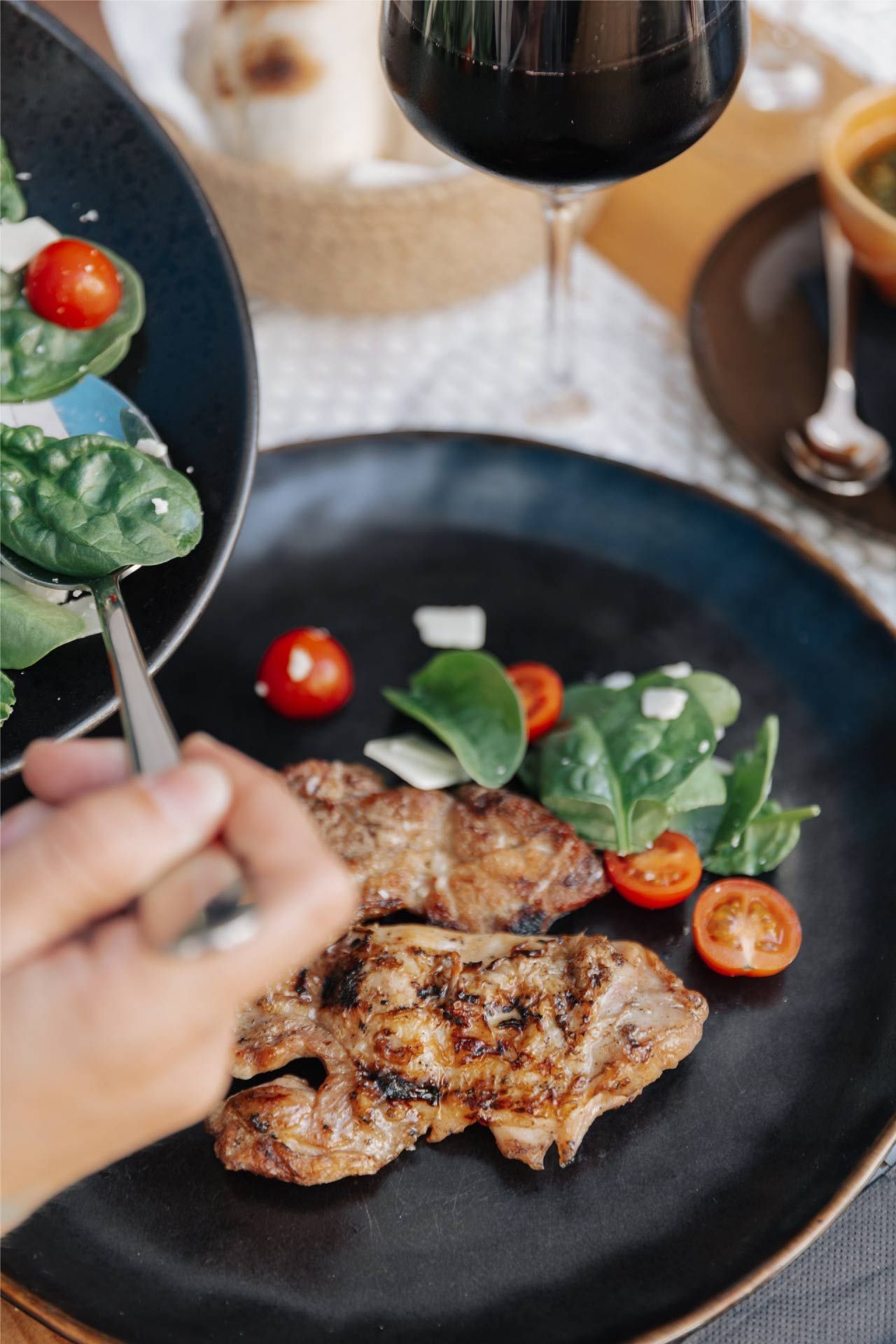 Persona sirviendo carne a la parrilla con ensalada y tomates en un plato negro, con una copa de vino tinto cerca.
