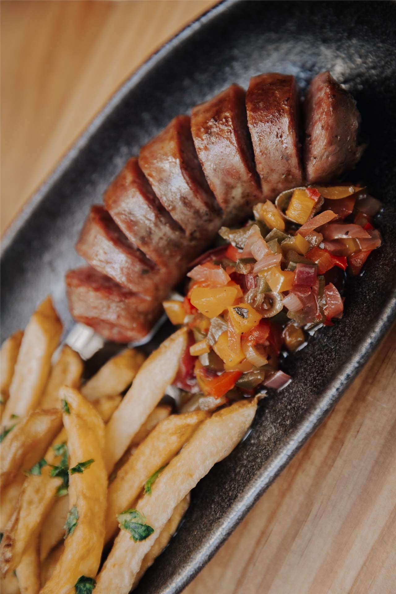 Salchicha, papas fritas y salsa en un plato negro. La salchicha está cortada en rodajas. Las papas fritas están doradas.