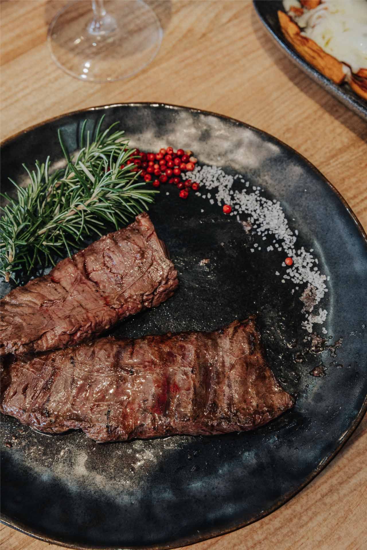 Bistec a la parrilla en un plato negro, adornado con romero, pimienta rosa y sal, sobre una mesa de madera.