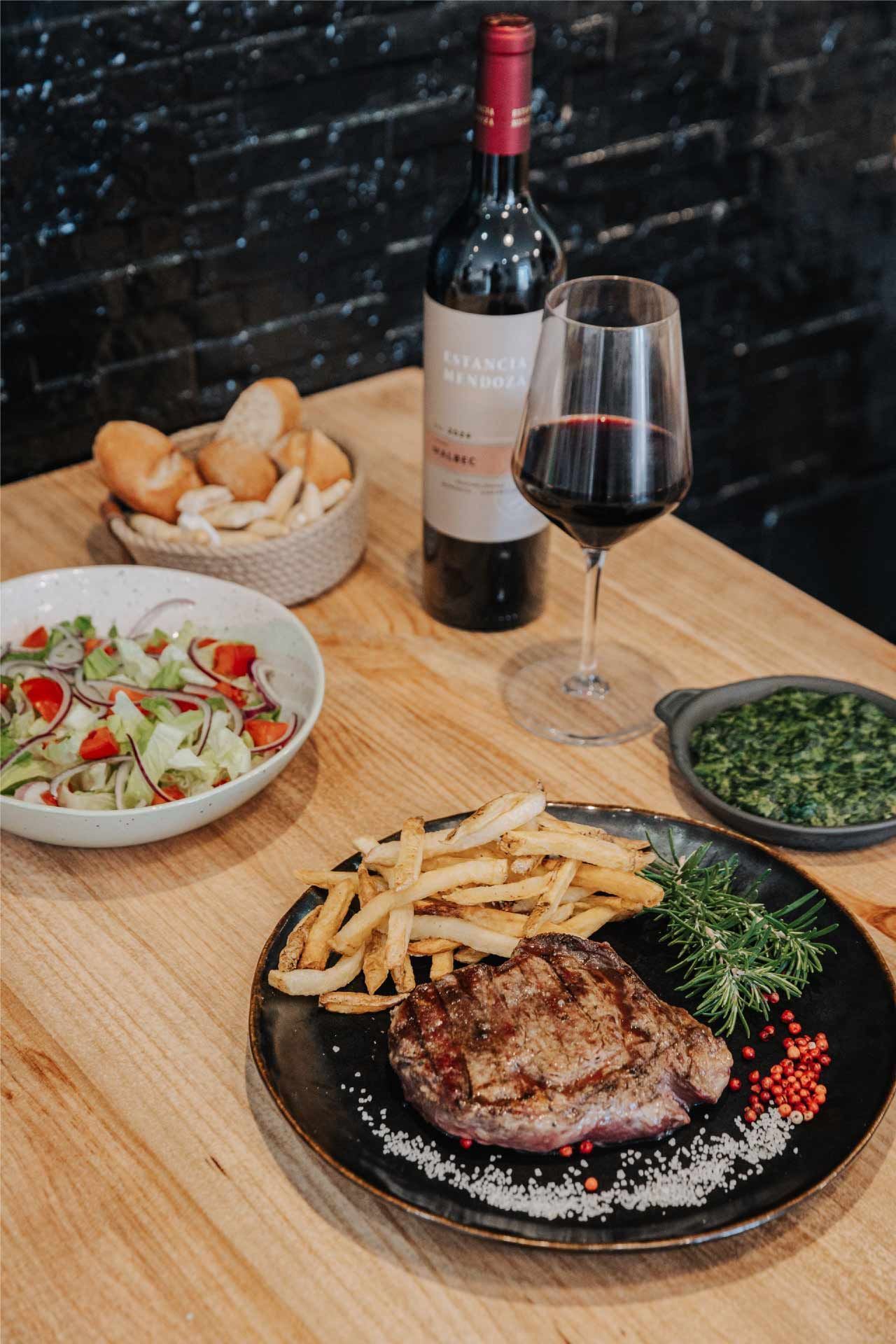 Cena de bistec: bistec a la parrilla, papas fritas, ensalada, vino tinto, pan y verduras en una mesa de madera.