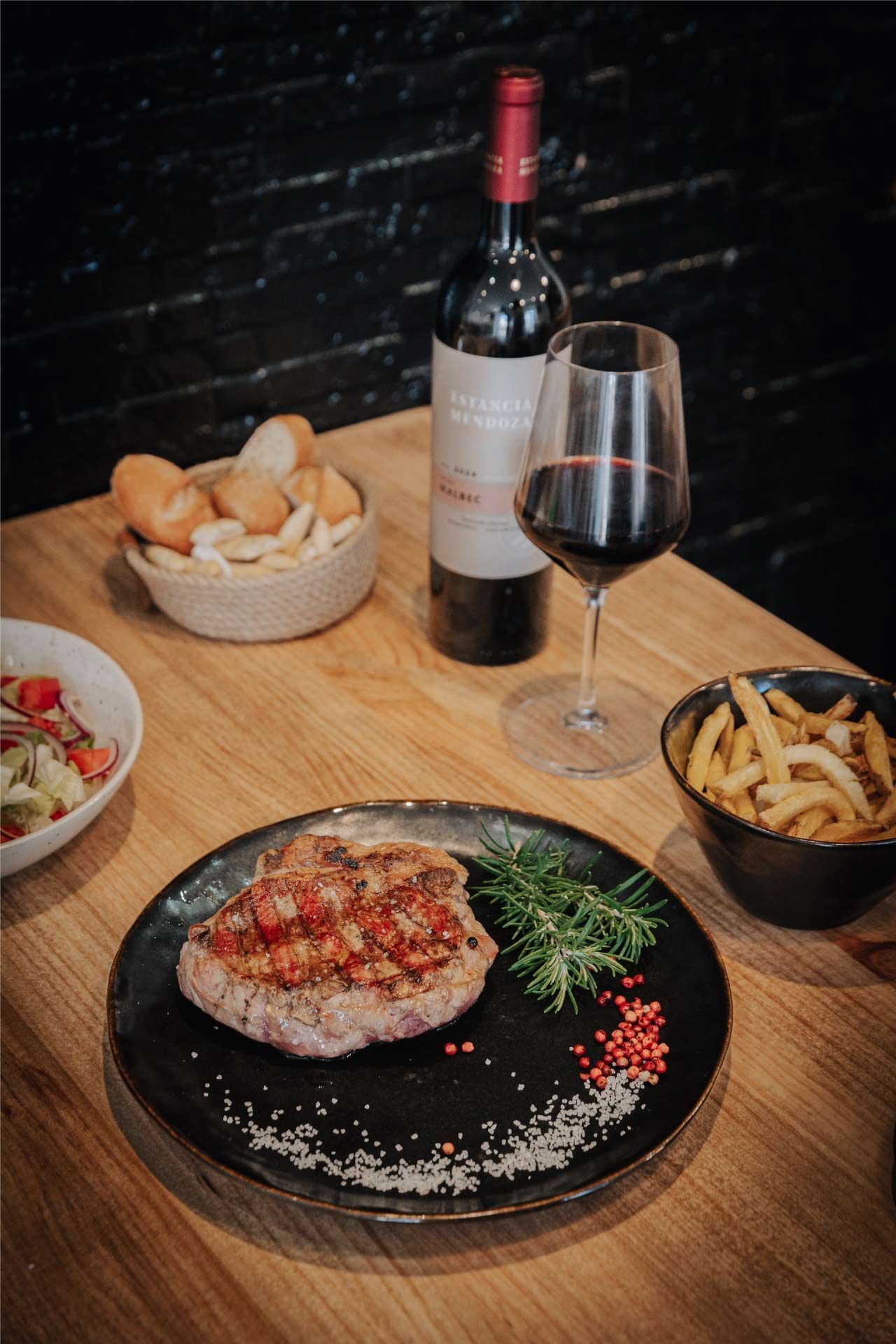 Bistec con aderezo rojo en un plato negro, vino, pan, papas fritas y ensalada en una mesa de madera.