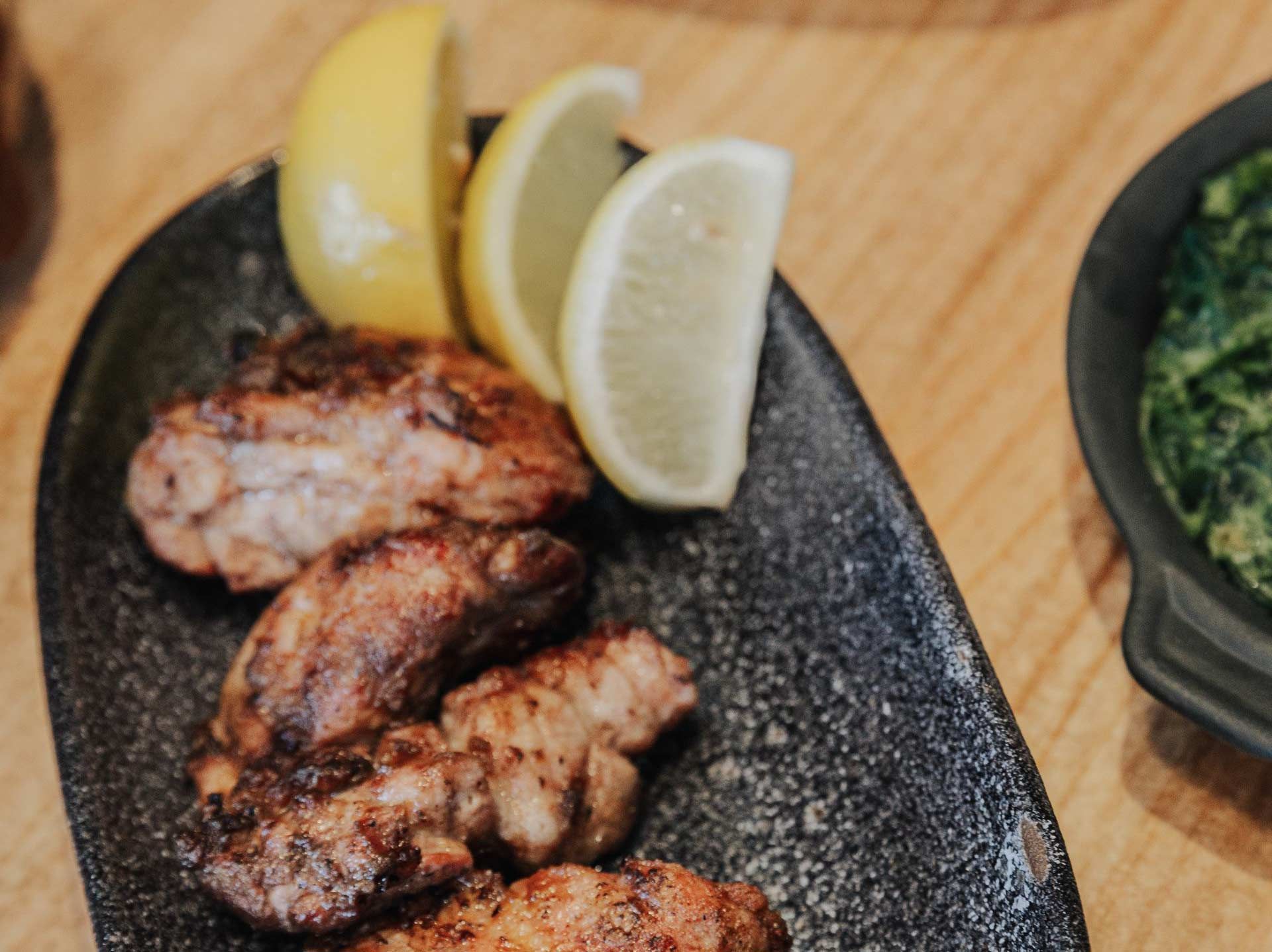 Alitas de pollo a la parrilla con rodajas de limón en un plato negro; guarnición de espinacas.