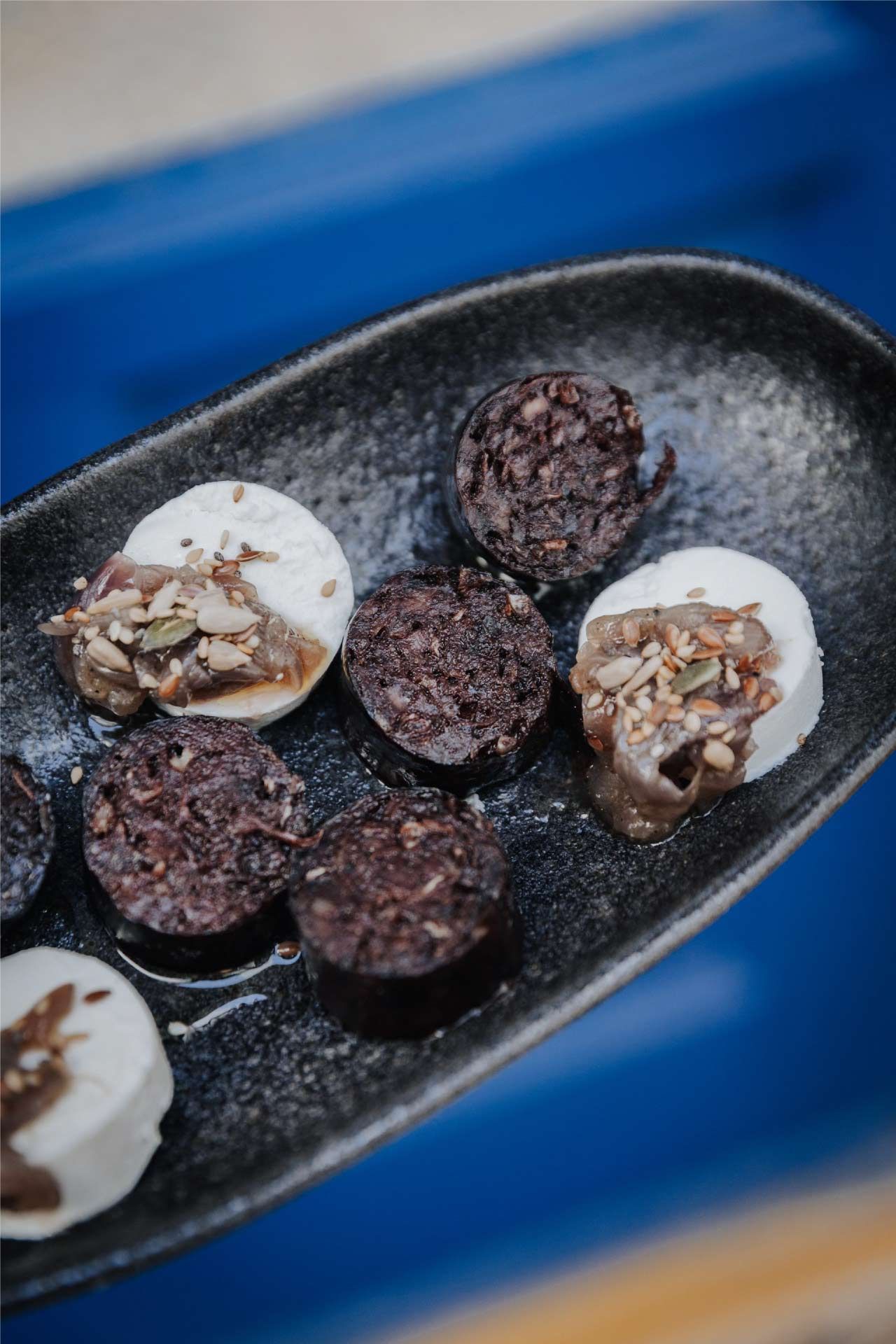 Plato negro con rodajas de queso de cabra y trozos oscuros tipo salchicha, fondo azul.
