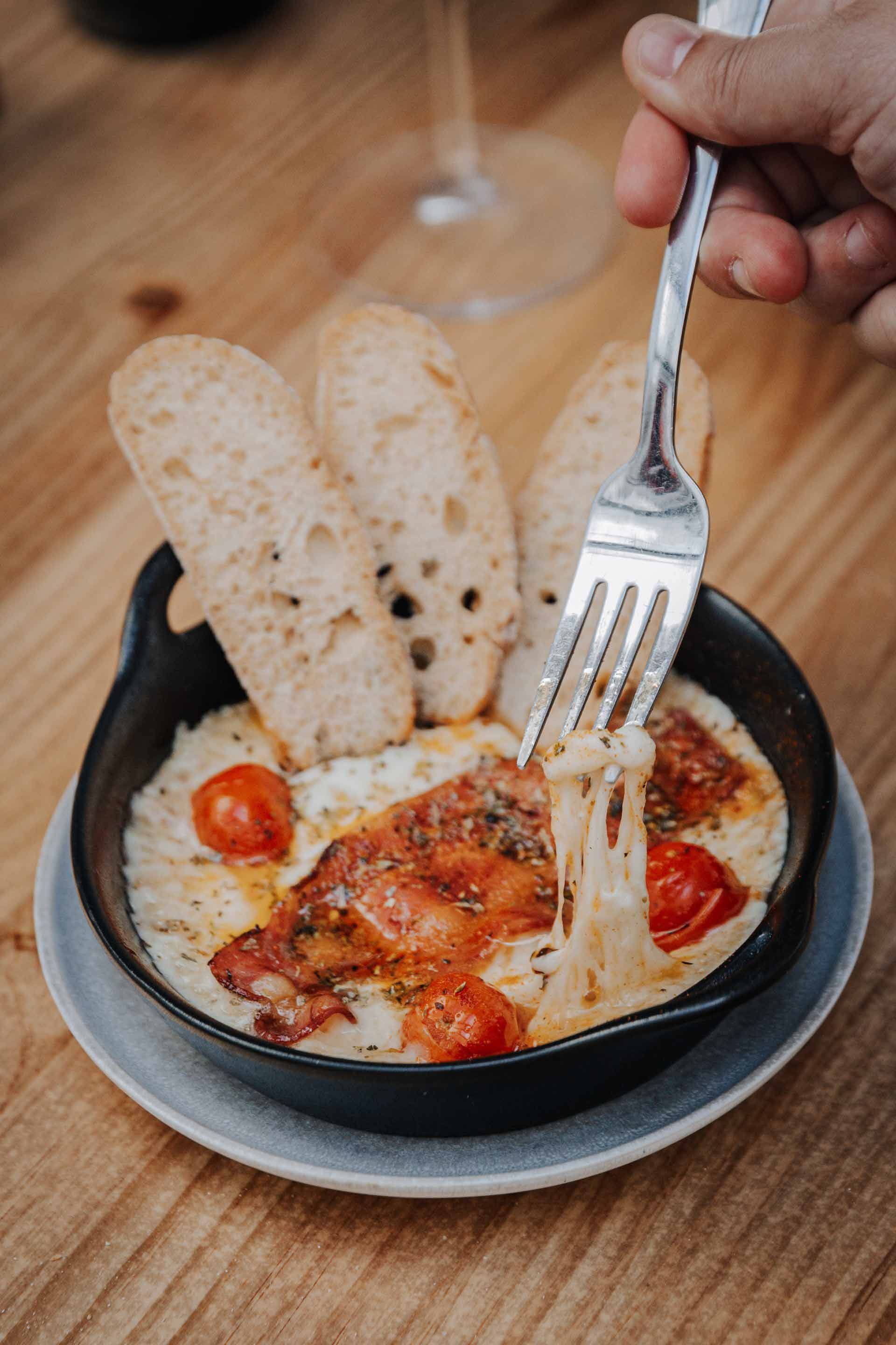 Mano usando un tenedor para sacar queso de una quesería horneada con pan y tomates cherry.