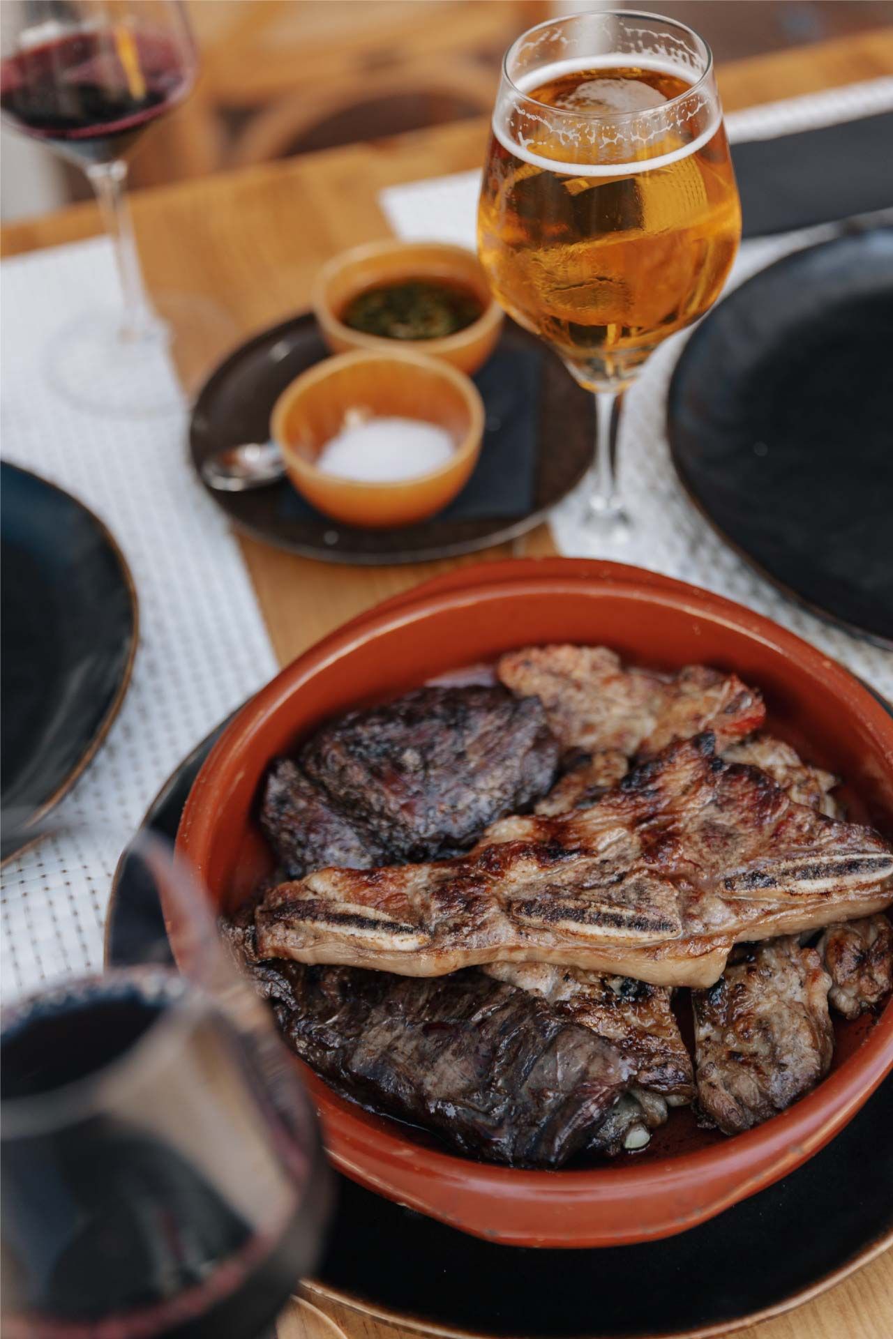 Costillas de res a la parrilla en una fuente de terracota con cerveza, vino tinto y salsas para mojar sobre una mesa de madera.