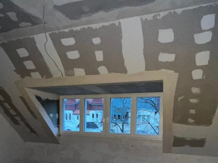 Innenansicht eines Dachzimmers mit Trockenbauwänden um ein Fenster, die den Blick auf Gebäude und Bäume einrahmen.